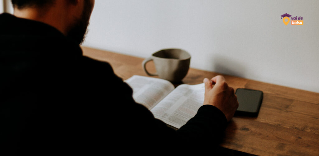 Imagem de um homem lendo um livro, com uma xícara de café e um smartphone sobre uma mesa de madeira, com o logo "Vai de Bolsa" no canto superior direito.