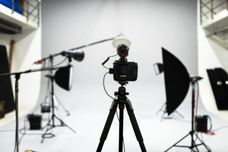 Imagem de um estúdio fotográfico profissional com uma câmera digital montada em um tripé no centro, cercada por diversos equipamentos de iluminação e softboxes, em um fundo branco.