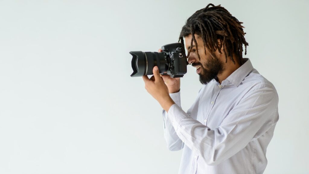 Imagem de um jovem fotógrafo negro, com cabelos em dreads, usando uma camisa xadrez branca e cinza, segurando uma câmera DSLR profissional. Ele sorri enquanto fotografa, ideal para conteúdo sobre faculdade de fotografia.