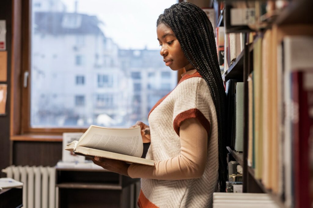 Imagem de uma jovem estudante negra com tranças, lendo um livro na biblioteca, ideal para ilustrar artigos sobre a faculdade de história. Ela está concentrada, em pé entre prateleiras de livros.