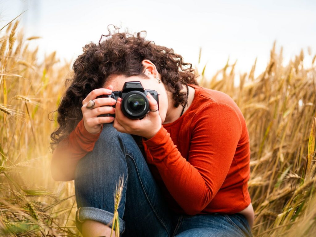 Imagem de uma fotógrafa com cabelos cacheados e camisa laranja, agachada em um campo de trigo, focando atentamente através de uma câmera DSLR. O cenário natural proporciona uma iluminação suave e agradável.